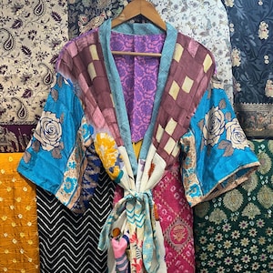 Könnte beinhalten: Ein farbenfroher Patchwork-Kimono-Mantel mit floralem und geometrischem Design. Der Mantel hat blaue, violette, rosa und gelbe Bahnen mit einem weißen Bindegürtel. Der Hintergrund zeigt verschiedene gemusterte Stoffe.