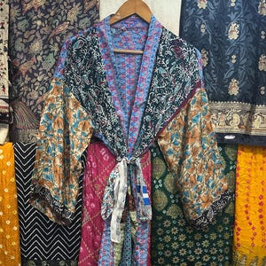 Peut inclure: Un kimono coloré et à motifs avec des manches longues est suspendu à un cintre en bois. La robe présente un mélange de motifs floraux et géométriques dans des tons de bleu, d'orange et de vert. Divers tissus à motifs sont visibles en arrière-plan.