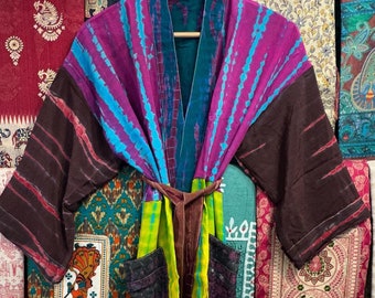 Puede incluir: Una bata kimono de patchwork colorida con un cinturón de atar. La bata está hecha de diferentes telas con varios patrones y colores, incluyendo morado, azul, verde, marrón y amarillo. La bata está colgada de una percha sobre un fondo de telas estampadas.