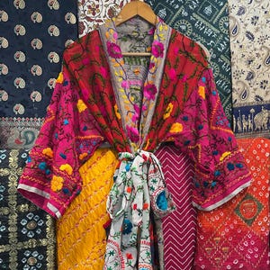 Könnte beinhalten: Ein lebendiger, bestickter Kimono in Pink- und Rottönen, verziert mit gelben und blauen Blumenakzenten. Der Kimono ist mit einem weißen Schal mit roter und grüner Stickerei gebunden. Verschiedene gemusterte Stoffe bilden den Hintergrund.