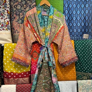 Op de afbeelding: Een kleurrijke, bedrukte kimono-badjas met een striksluiting hangt aan een houten hanger. De badjas heeft paisley-, bloemen- en geometrische ontwerpen in oranje, groen, blauw en rood. Verschillende stoffen rollen in verschillende kleuren en patronen zijn op de achtergrond.