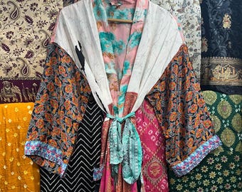 Może przedstawiać: Kolorowe, wzorzyste kimono z wiązaniem, wiszące na drewnianym wieszaku. Szlafrok ma mieszankę kwiatowych, paisley i geometrycznych wzorów w odcieniach pomarańczu, różu, błękitu i bieli. Tło przedstawia różne wzory tkanin.