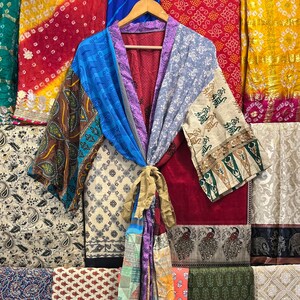 May include: A patchwork kimono robe in shades of blue, purple, beige, and brown hangs on a wooden hanger. The robe is tied with a gold-colored sash. Various colorful fabrics are visible in the background, including red, yellow, and green.
