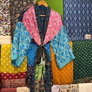 May include: A colorful patchwork kimono robe with a floral and paisley pattern. The robe features shades of pink, blue, green, and black, with a black tie closure. The robe is hanging on a wooden hanger against a backdrop of patterned fabrics.