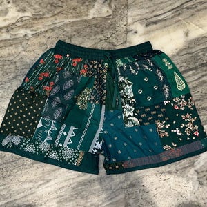 Puede incluir: Shorts patchwork verdes con cintura con cordón. Los shorts presentan varios patrones y diseños florales en tonos verdes, blancos y rojos. Los shorts están hechos de una tela ligera.