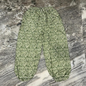 May include: Green patterned trousers with a drawstring waist and elasticated cuffs. The trousers feature a repeating floral design in shades of green. The fabric appears lightweight and suitable for casual wear.
