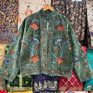 Op de afbeelding: Een groen fluwelen jasje met bloemenborduursel. Het jasje heeft een mandarijnsluiting en een voorsluiting. Het borduursel is in tinten blauw, rood en oranje.