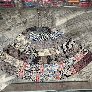Peut inclure: Une robe d'été patchwork avec un corsage à motifs et une jupe à volants. La jupe présente une variété de motifs de tissu dans les tons de bleu, noir, marron et crème. La robe est présentée sur une surface en marbre.