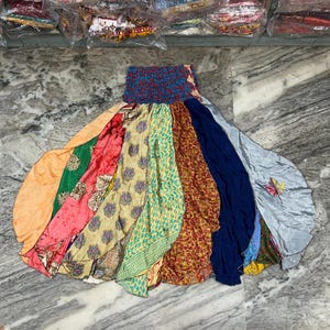 May include: A colorful, bohemian-style skirt with a smocked waistband. The skirt features panels of various fabrics in shades of orange, pink, green, yellow, blue, and red. The skirt is laid out on a marble surface.
