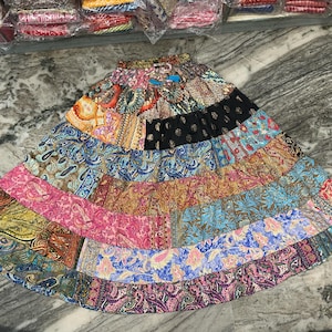 May include: A vibrant patchwork skirt with a flared silhouette. The skirt showcases a mix of patterned fabrics in blue, pink, orange, and black. It has an elasticated waistband and is likely made from a lightweight material, suitable for warm weather.