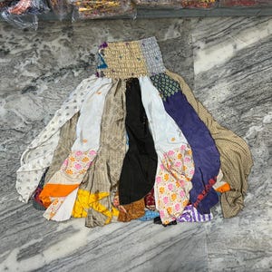 May include: A patchwork skirt with a gathered waistband. The skirt is made from panels of different fabrics in white, black, beige, orange, purple, and floral prints. The skirt is displayed on a marble surface.
