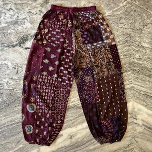 May include: Burgundy patchwork trousers with an elasticated waistband. The trousers feature various patterned fabrics in shades of burgundy, brown, and purple. The design includes floral, geometric, and paisley motifs, creating a bohemian style.