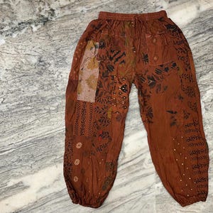 Peut inclure: Pantalon marron avec un motif patchwork composé de motifs floraux et géométriques. Le pantalon a une taille à cordon et une coupe ample.