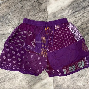 Puede incluir: Pantalones cortos morados con patchwork, cintura elástica y cordón. Los pantalones cortos presentan varios paneles de tela estampada en tonos morados, blancos y dorados. Ropa informal.