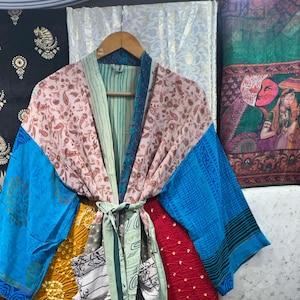 Könnte beinhalten: Ein farbenfroher Kimono-Mantel mit Bindegürtel. Der Mantel zeigt eine Mischung aus Blumen-, Paisley- und geometrischen Mustern in Rosa-, Blau- und Grüntönen. Der Kimono hängt an einem Holzbügel.