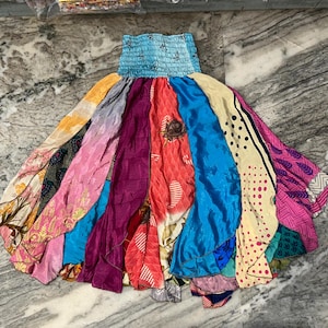 May include: A colorful, patchwork skirt with a light blue, elasticized waistband. The skirt features a variety of fabrics in shades of pink, purple, blue, yellow, and orange. The skirt's design is a flowing, layered style.