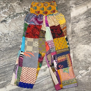 Può includere: Pantaloni patchwork con elastico giallo in vita e una varietà di toppe in tessuto colorato. Le toppe includono motivi floreali, geometrici e astratti nelle tonalità del rosso, blu, verde e giallo. I pantaloni sono realizzati in un materiale leggero.