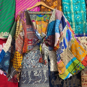 Op de afbeelding: Een kleurrijk patchwork top gemaakt van hergebruikte stof. De top heeft een strik aan de voorkant en lange mouwen. De stof is een mix van verschillende patronen en kleuren, waaronder bloemen, geometrisch en paisley.