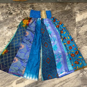 May include: Wide-leg trousers in a patchwork design, featuring various shades of blue and patterned fabrics. The waistband is solid blue, with a gold accent. The trousers are designed for a relaxed fit and are made from a variety of fabrics.