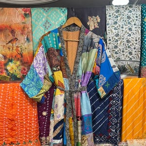 Puede incluir: Una bata kimono patchwork con una variedad de patrones de tela coloridos, colgada de una percha de madera. La bata presenta una mezcla de azules, verdes, amarillos y rojos. Varias telas estampadas se exhiben detrás de la bata.