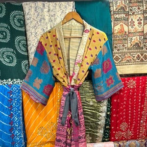 May include: A colorful, patterned kimono robe with a tie closure hangs on a wooden hanger. The robe features a mix of yellow, blue, and pink fabrics. The background displays various textiles in different colors and patterns.