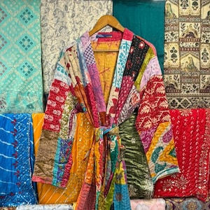 May include: A long-sleeved robe featuring a patchwork design made from various colorful fabrics with intricate patterns. The garment includes a matching fabric tie belt at the waist and is constructed with visible quilted stitching throughout the entire piece.