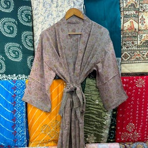 May include: A light purple floral print robe with a matching tie, hanging on a wooden hanger. The robe is displayed in front of a variety of colourful fabrics with different patterns and designs. The robe is made of a lightweight material.