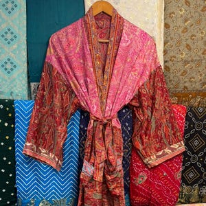 Puede incluir: Una bata tipo kimono de manga larga que presenta una mezcla vibrante de tela rosa y rojo intenso con un intrincado patrón de cachemira. La prenda incluye un cinturón de tela a juego en la cintura y mangas anchas, colgada de una percha de madera.
