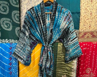Puede incluir: Una bata con estampado tie-dye azul, negro y blanco cuelga de una percha de madera. La bata se exhibe sobre un fondo de varias telas coloridas, incluyendo estampados rojos, amarillos y florales. La bata está atada a la cintura con un cinturón de tela a juego.