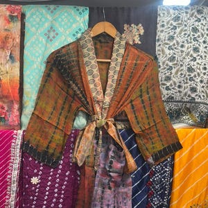 May include: A tie-dye kimono robe in shades of orange and brown, with a patterned collar and tie, displayed on a wooden hanger. The robe is surrounded by a variety of colourful fabrics with diverse patterns, including floral and geometric designs.