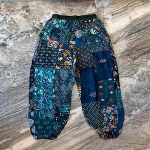 Op de afbeelding: Losvallende broek met een patchworkontwerp bestaande uit diverse blauwe en groene stoffen panelen. Het kledingstuk toont een mix van bloemen-, paisley- en geometrische patronen over het oppervlak, met een elastische tailleband aan de bovenkant.