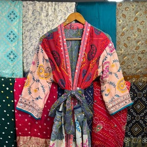 May include: A long-sleeved robe featuring a patchwork design with red, peach, and white panels. The garment is decorated with colourful floral embroidery, small circular mirror accents, and a matching fabric tie belt at the waist.