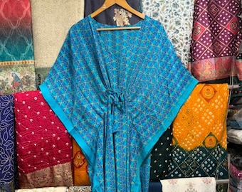 May include: A turquoise blue kaftan dress with a geometric pattern, hanging on a wooden hanger. The dress has a tie closure at the waist and is surrounded by colorful fabrics. The kaftan is a loose-fitting garment.