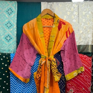 Peut inclure: Un peignoir de style kimono à manches longues présentant un motif patchwork vibrant avec des panneaux orange vif, roses et jaunes. Le vêtement comprend une ceinture à nouer assortie à la taille et un large col, mélangeant des motifs floraux et animaliers.