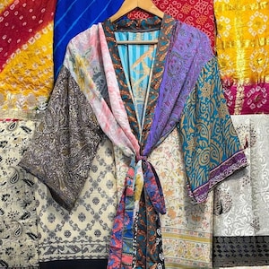 May include: A colorful patchwork silk robe with a tie belt. The robe is made of different fabrics with various patterns and colors, including pink, purple, blue, and green. The robe is hanging on a hanger against a background of colorful fabrics.