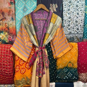 Op de afbeelding: Een kleurrijke kimono-achtige robe met een strik in de taille. De robe heeft een mix van oranje, gele, paarse en gouden stoffen. De achtergrond toont verschillende textielsoorten in blauw, rood en groen.