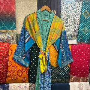 Pode incluir: Um roupão quimono colorido em estilo patchwork com mangas compridas, pendurado em um cabide de madeira. O roupão apresenta uma mistura de tecidos azuis, amarelos, verdes e estampados. Vários tecidos coloridos são visíveis no fundo.