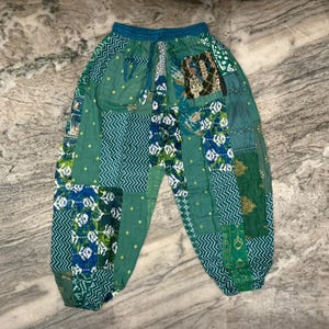 Pode incluir: Calças verdes de patchwork com cintura com cordão. As calças apresentam várias tonalidades de verde, azul e branco num design de patchwork. O tecido parece leve, adequado para roupa casual.
