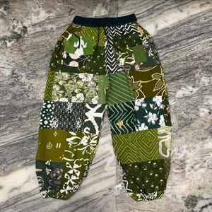 Pode incluir: Calças de patchwork com uma variedade de quadrados de tecido verdes e brancos com diversos padrões florais, geométricos e abstratos. As calças têm uma cintura elástica de cor escura e punhos elásticos nos tornozelos.