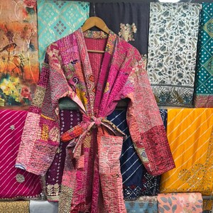 Könnte beinhalten: Ein Patchwork-Bademantel in Rosa-, Rot- und Lilatönen hängt an einem Holzbügel. Der Bademantel hat einen Bindverschluss und ist von bunten Stoffen mit verschiedenen Mustern und Designs umgeben.