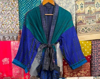 Puede incluir: Una bata estilo kimono de manga larga con un diseño de bloques de color en azul verdoso y azul real. La prenda incluye un cinturón de lazo gris oscuro y un cuello gris oscuro, con sutiles patrones verticales de teñido anudado en la tela.