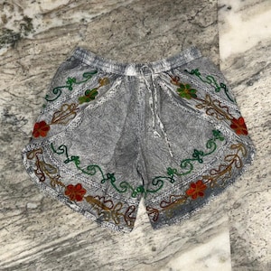 May include: Gray shorts with an embroidered floral design in red, green, and brown. The shorts have a drawstring waist and are made of a textured fabric. The floral embroidery is along the bottom edge of the shorts.