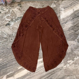 May include: A pair of brown wrap-style pants featuring a high-waisted design with a lace-up front closure. The garment has wide, overlapping leg panels that are decorated with intricate, dark-toned embroidery along the curved bottom edges.