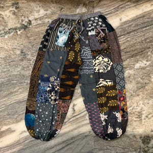May include: Patchwork trousers featuring a variety of dark-toned fabric panels in shades of black, grey, brown, and blue. The garment includes an elastic waistband with a drawstring, cuffed ankles, and a mix of floral, geometric, and paisley patterns.