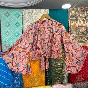 May include: A pink paisley print wrap top with wide sleeves, tied at the waist. The top is displayed on a wooden hanger against a backdrop of colorful fabrics. The top features a mix of orange, blue, and red accents.