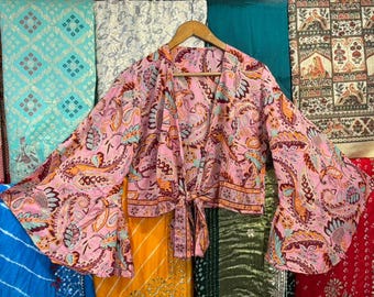 May include: A pink paisley print wrap top with wide sleeves, tied at the waist. The top is displayed on a wooden hanger against a backdrop of colorful fabrics. The top features a mix of orange, blue, and red accents.