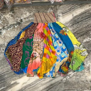 Op de afbeelding: Een lange, uitlopende rok met een brede, gesmokte elastische tailleband. De rok is opgebouwd uit meerdere verticale stroken stof, elk met een eigen patroon, waaronder bloemenprints, geometrische vormen en wervelingen in levendige kleuren.