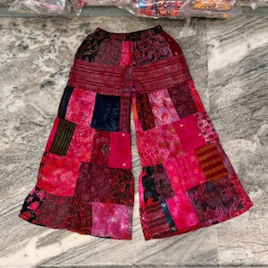Op de afbeelding: Wijd uitlopende patchworkbroek in roze, rode en bordeaux tinten. De broek heeft een verscheidenheid aan stofpatronen en texturen, waardoor een bohemien stijl ontstaat. De tailleband is effen bordeaux van kleur.