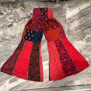 Op de afbeelding: Patchwork palazzo broek in tinten rood, bordeaux en marineblauw. De broek met wijde pijpen heeft een verscheidenheid aan stoffen panelen met patronen, waardoor een bohemian stijl ontstaat. De tailleband is elastisch. De broek ligt plat op een marmeren oppervlak.