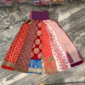 Puede incluir: Un pantalón patchwork con diseño de pernera ancha. El pantalón presenta una variedad de tejidos coloridos, incluyendo rojo, marrón y rosa, con diferentes patrones y texturas. La cintura es de material elástico morado oscuro.